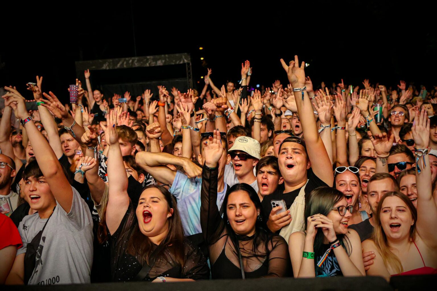 Large concert crowd with raised hands and phone lights in dark venue